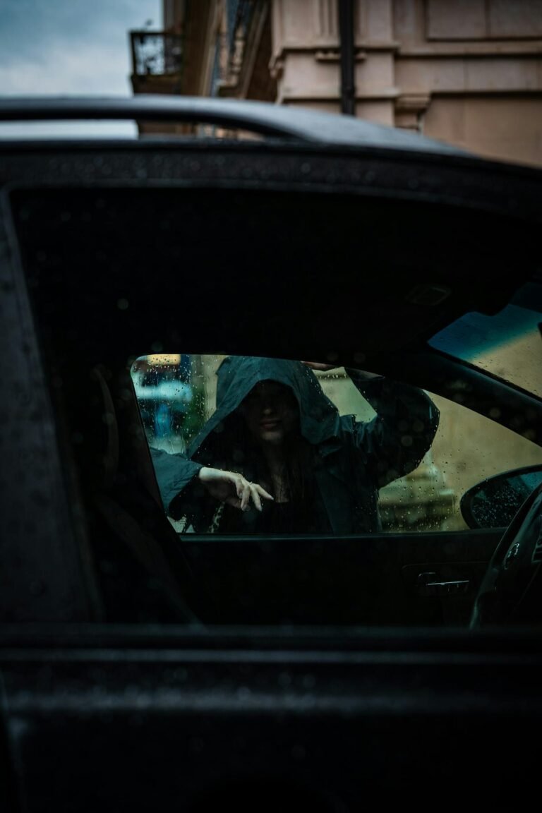 Dark, rainy scene with a hooded figure outside a car window.